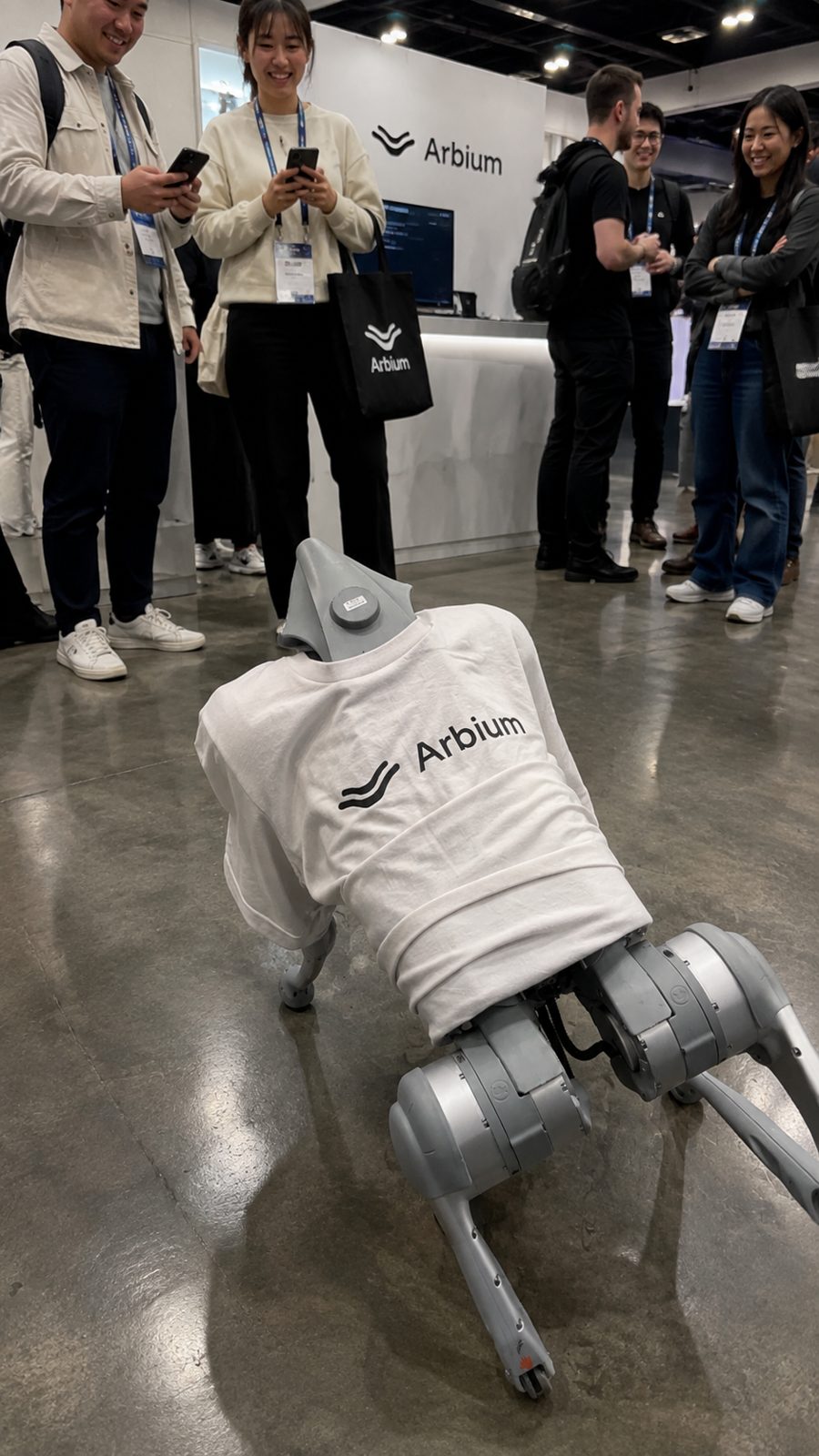 Robot dog in an Arbium shirt with attendees around a booth.