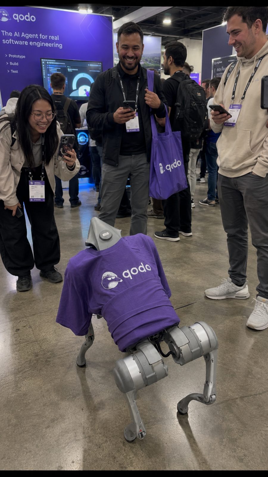 People filming a robot dog in a Qodo shirt on a San Francisco conference floor.